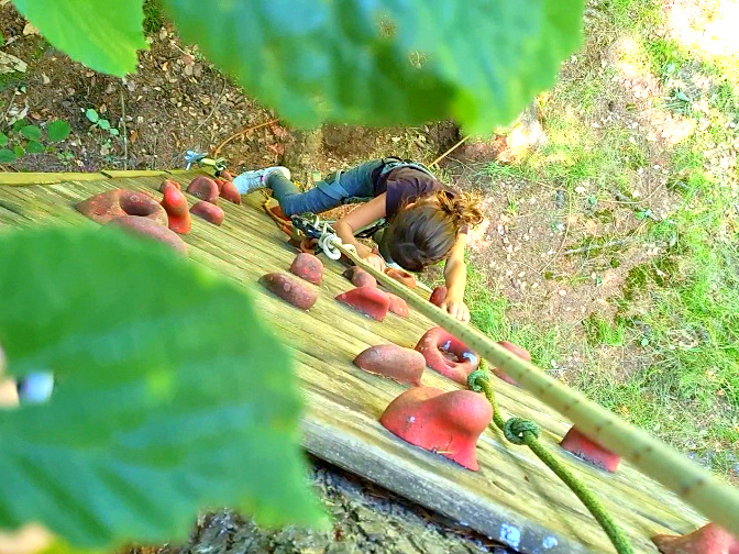 Jeune fille escaladant le mur d'escalade du premier atelier du deuxième parcours d'accrobranche au camping Les Trois Sources, activité nature et familiale.