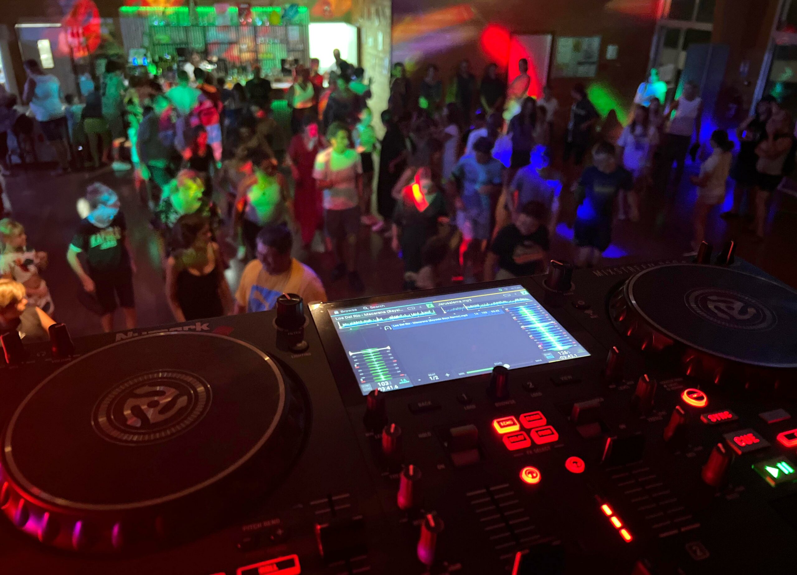 Point de vue du DJ avec platines et foule dansant dans la salle polyvalente du camping Les Trois Sources, ambiance musicale et événement familial.