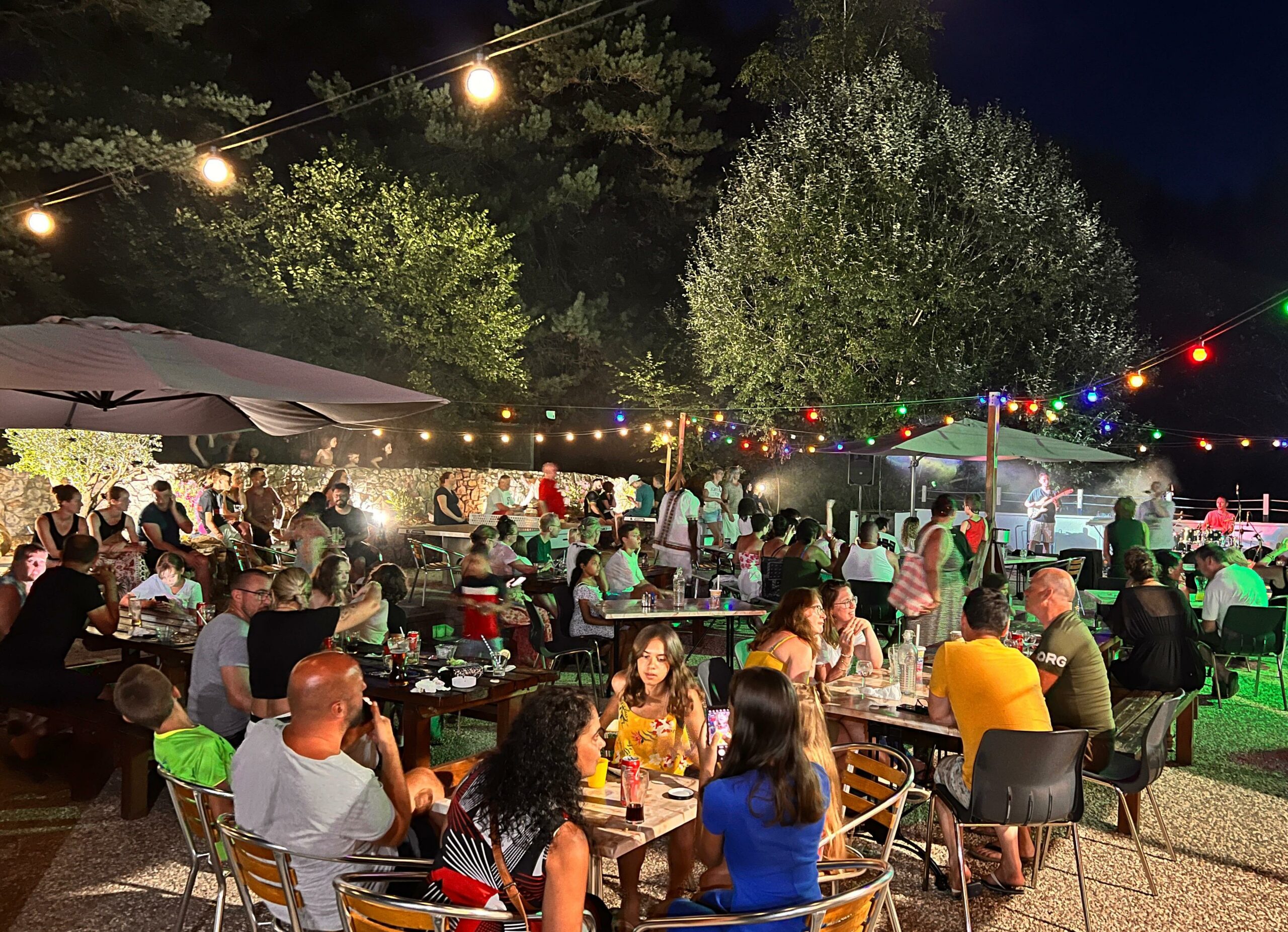 Personnes à table sur la terrasse du restaurant du camping Les Trois Sources assistant à un concert de nuit sous des guirlandes lumineuses, ambiance conviviale et familiale en plein air.