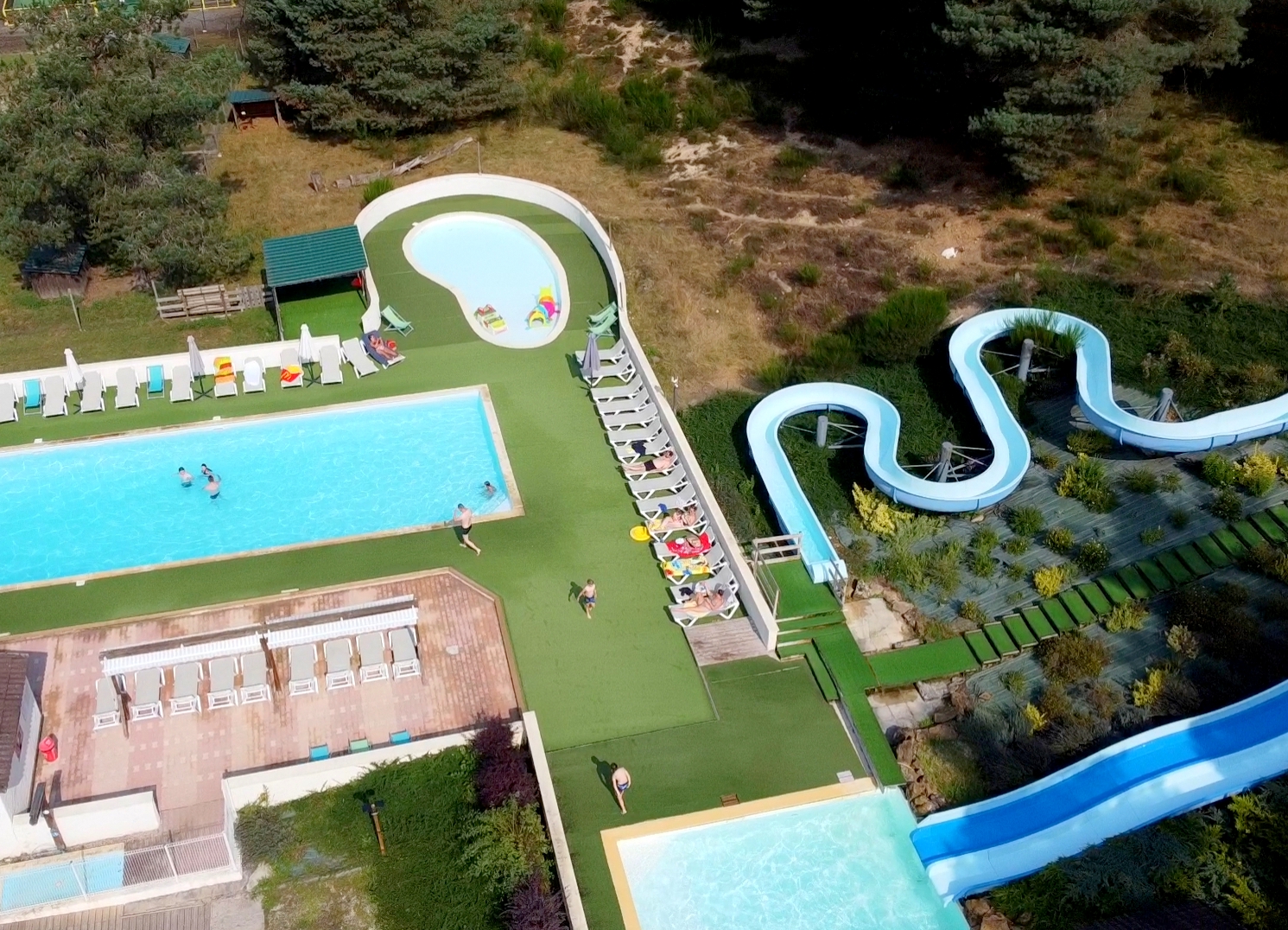 Vue aérienne du parc aquatique du camping Les Trois Sources avec grand bassin, pataugeoire, toboggans et transats, espace détente et loisirs en famille au cœur de la nature.